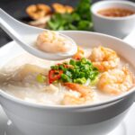 白色瓷碗中盛著熱氣騰騰的海鮮廣東粥，蝦仁、干貝與魚片鋪在上層，粥質細膩綿滑，湯勺微掀起粥面