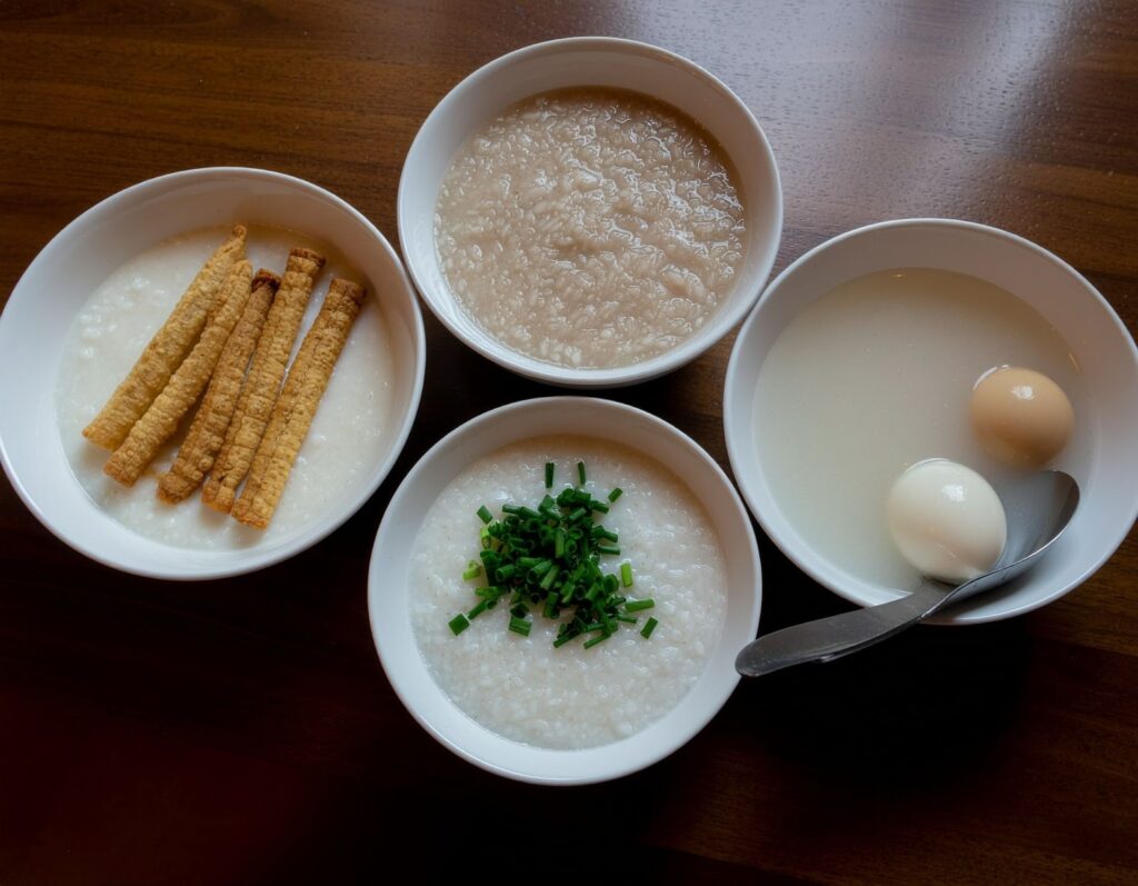 餐桌上放著三碗質地不同的廣東粥，旁邊有蔥花、油條與皮蛋等配料，展現不同米種煮出的濃稠度與風味對比。